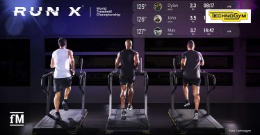 Drei Personen laufen auf Laufbändern in einem dunklen Raum, Blick von hinten, Displays vor ihnen, darüber eingeblendetes Leaderboard mit Namen Andrew, Dylan, John und Max, Distanzen und Zeiten.
