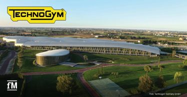 Luftaufnahme eines großen modernen Gebäudekomplexes mit geschwungenem Dach in grüner Landschaft, links rundes Gebäude, im Vordergrund Wege und Basketballfeld, oben links gelbes Logo „Technogym“, Umgebung mit Feldern und Siedlungen.