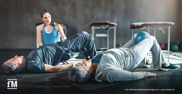 Im Vordergrund liegen ein älterer Mann und eine ältere Frau auf Trainingsmatten und führen eine Brückenübung aus. Hinter ihnen sitzt eine Trainerin im blauen Oberteil auf dem Boden und beobachtet die Übungsausführung. Im Hintergrund stehen Trainingsliegen und Gymnastikbälle