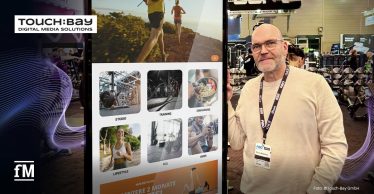 Digitale Stele mit TOUCH:BAY-Benutzeroberfläche neben einer Person in heller Kleidung in einer belebten Fitnessmessehalle.