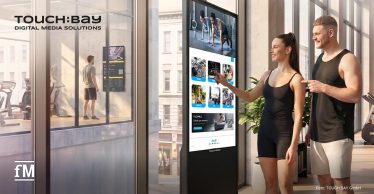 Im Vordergrund bedienen eine Frau und ein Mann in Sportkleidung einen freistehenden digitalen TOUCH:BAY Bildschirm. Auf dem Display sind mehrere Kacheln mit Trainings- und Lifestyleinhalten sichtbar. Links hinter einer Glasfront steht eine Person vor einem weiteren Display. Oben links steht das Logo „TOUCH:BAY DIGITAL MEDIA SOLUTIONS“