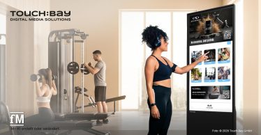 Frau mit Kopfhörern bedient ein großes TOUCH:BAY-Display neben Trainingsgeräten in einem Fitnessstudio.