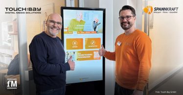 Zwei Männer links und rechts neben einer freistehenden digitalen Stele, beide mit erhobenem Daumen; mittig ein großer Touchscreen mit Benutzeroberfläche „Körperanalyse“, Buttons „Anfänger“ und „Fortgeschrittene“, QR-Code unten rechts; links Logo „TOUCH:BAY Digital Media Solutions“, rechts Logo „Spannkraft Rehasport Fitness Gesundheit“