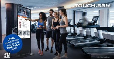 Vier Personen vor Touchscreen-Stelen im Fitnessstudio, rechts mehrere Laufbänder, Schriftzug „TOUCH:BAY“ oben rechts.