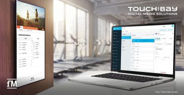 Vordergrund rechts ein Laptop mit Magicline Backend-Ansicht, links ein stehendes TOUCH:BAY-Display mit Websitebereich „AKTUELLER KURSPLAN“, Navigationspunkten „STUDIO“, „TRAINING“, „ERNÄHRUNG“, „LIFESTYLE“, „FAQ“, „NEWS“ und Logo „BRANDNAME“ unten; auf dem Laptop erscheinen unter anderem „Kurse“, „Kursplan“, „Zumba“, „Verwaltung“, „Teilnehmer“, „Historie“, „KURSTERMINE PLANEN“, „TERMIN BUCHEN“ und „KURS LÖSCHEN“ vor unscharfem Fitnessstudiohintergrund.