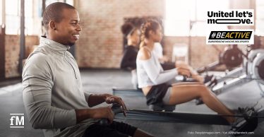 Mehrere Personen trainieren gemeinsam an Ruderergometern in einem lichtdurchfluteten Fitnessstudio mit Backsteinwänden. Im Fokus steht ein junger Mann in hellem Sportoberteil, der motiviert und lächelnd trainiert. Rechts oben ist das Logo der Kampagne '#BEACTIVE – Europäische Woche des Sports' und 'United lte's move!' zu sehen.