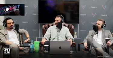 Drei Männer sitzen in Sesseln vor Mikrofonen und nehmen einen Podcast auf. In der Mitte sitzt Janosch Marx, trägt ein helles Hemd und hat ein MacBook vor sich. Links sitzt ein Mann mit beiger Jacke, weißem T-Shirt und orangefarbenen Sonnenbrillen, rechts ein Mann in grauem Anzug mit weißem Shirt. Alle tragen große Kopfhörer. Im Hintergrund sind TRINITY-Logos auf grauem Stoff und ein ausgeschalteter Flachbildschirm zu sehen. Links oben ist das farbige UPSHIFT-Logo eingeblendet, unten rechts steht der Fotohinweis: 'Foto: TRINITY Digital'. Ein Getränkebecher mit Strohhalm und eine Wasserflasche stehen auf dem Tisch.