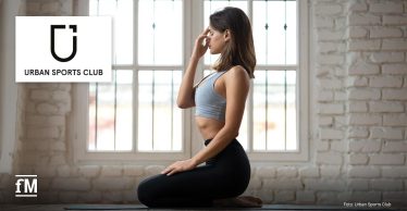 Frau im Sportoutfit sitzt auf Yogamatte und übt Atemtechnik in hellem Raum. Links oben ist das Logo von Urban Sports Club zu sehen.