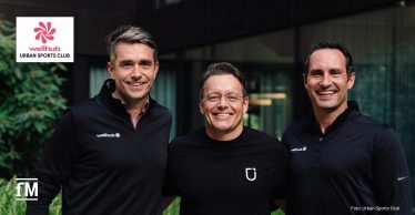 Drei lächelnde Männer stehen eng nebeneinander in einem begrünten Innenhof vor einer modernen Glasfassade. Alle tragen dunkle Oberteile, die beiden äußeren Männer tragen blaue Jeans.