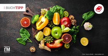 Draufsicht auf dunkle Tischoberfläche mit Holzbrett, darauf Brokkoli, Paprika, Zitrusfrüchte, Apfel, Kiwi, Spinat, Ingwer und Nüsse verteilt.