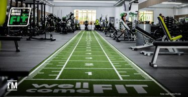 Trainingsbereich in einem Fitnessstudio mit grünem Kunstrasen, Kraftgeräten, Hantelbänken und mehreren Trainingsmaschinen in einer großen Halle.
