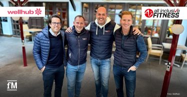 Vier lächelnde Vertreter von Wellhub und all inclusive Fitness stehen Arm in Arm unter einem Pavillon und tragen sportlich-legere Kleidung. Im Hintergrund sind die Logos der beiden Unternehmen zu sehen.