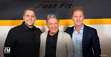 Drei Männer (v.l. Eric Krahnert (Wellhub), Frank Böhme (Just Fit) und Samuel Turnwald (Wellhub)) stehen nebeneinander vor einer Wand mit dem Just Fit-Logo.