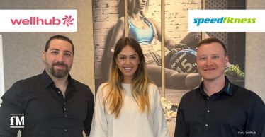 Drei lächelnde Personen stehen in einem modern eingerichteten Raum vor einem großformatigen Wandbild mit Fitnessmotiv.