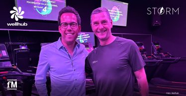 Zwei Personen stehen in einem STORM Fitnessstudio vor Laufbändern, im Hintergrund sind Bildschirme mit Trainingsinformationen zu sehen. Oben links ist das Wellhub-Logo, oben rechts das von STORM und unten links das fM-Signet, alles in Weiß.