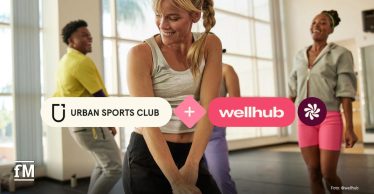 Vier Menschen trainieren in heller Fitnesshalle, im Vordergrund eine Frau im grauen Top. Logos von Urban Sports Club und Wellhub sind eingeblendet.