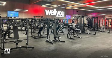 Innenraum eines Fitnessstudios in Hückelhoven, aufgenommen aus zentraler Perspektive mit Blick in die Trainingsfläche; im Vordergrund mehrere schwarze Kraftgeräte mit Sitzpolstern und Metallrahmen, im Mittelbereich weitere Geräte und Kabelzüge, im Hintergrund eine Wand mit beleuchtetem „wellyou“-Schriftzug
