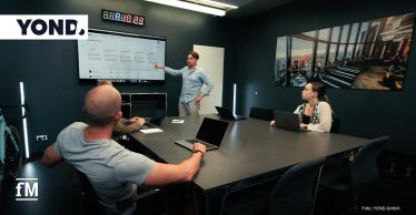 Konferenzraum mit dunkelblauen Wänden: Ein Mann steht neben einem großen Wandmonitor, zeigt auf Diagramme des YOND‑Dashboards; drei sitzende Personen schauen zu, vor ihnen Laptops auf einem großen Tisch; links ein Rennrad an der Wand.
