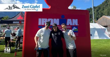 Drei Männer stehen vor einer großen roten Ironman-Figur auf einer Wiese, im Hintergrund Zelte, Berge und blauer Himmel